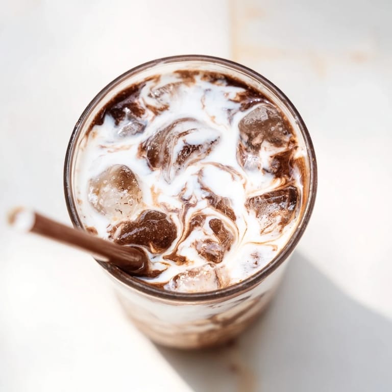 Stirring the chocolatey Milo Ice Latte Hack in a glass, with a spoon and milk nearby for a refreshing treat.